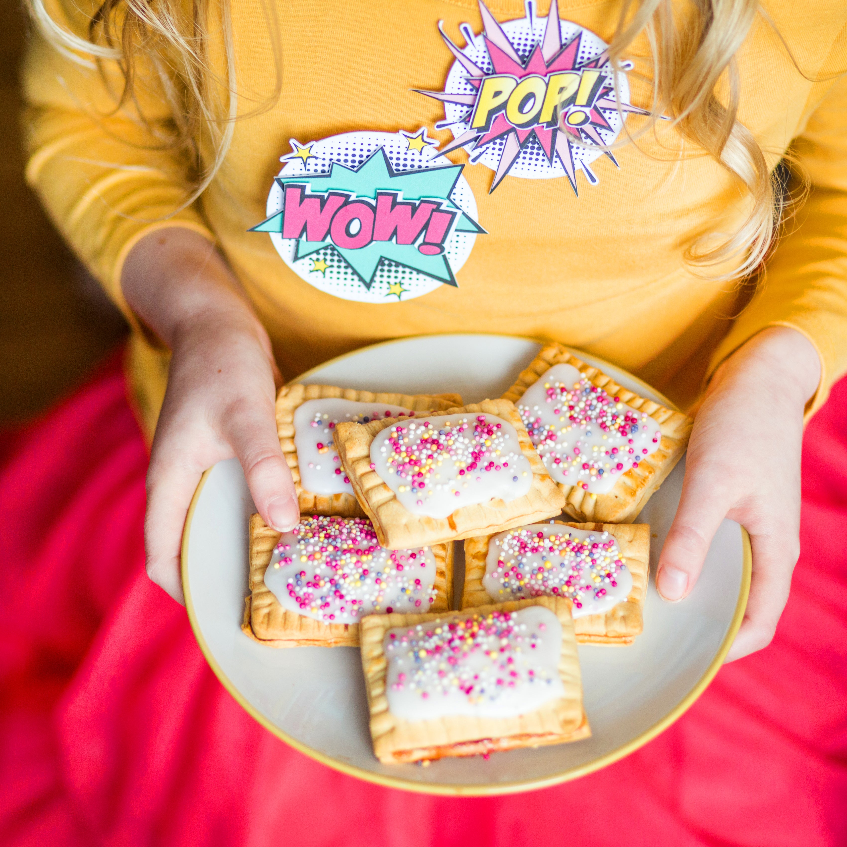 Child holding a plate of frosted pastries with colorful sprinkles, wearing a yellow shirt and pink skirt with comic-style stickers.