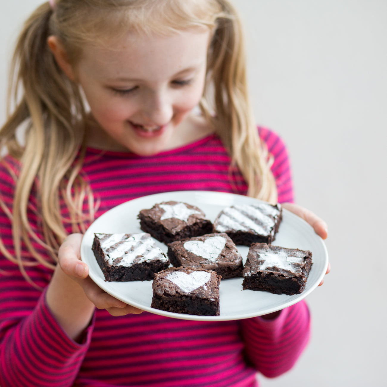 Child in a pink striped shirt holding a plate of heart-patterned brownies, smiling down at them.