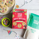 Craft & Crumb Dotty Biscuits kit with colorful candies, biscuit mix, and decorated cookies on a light wooden surface.