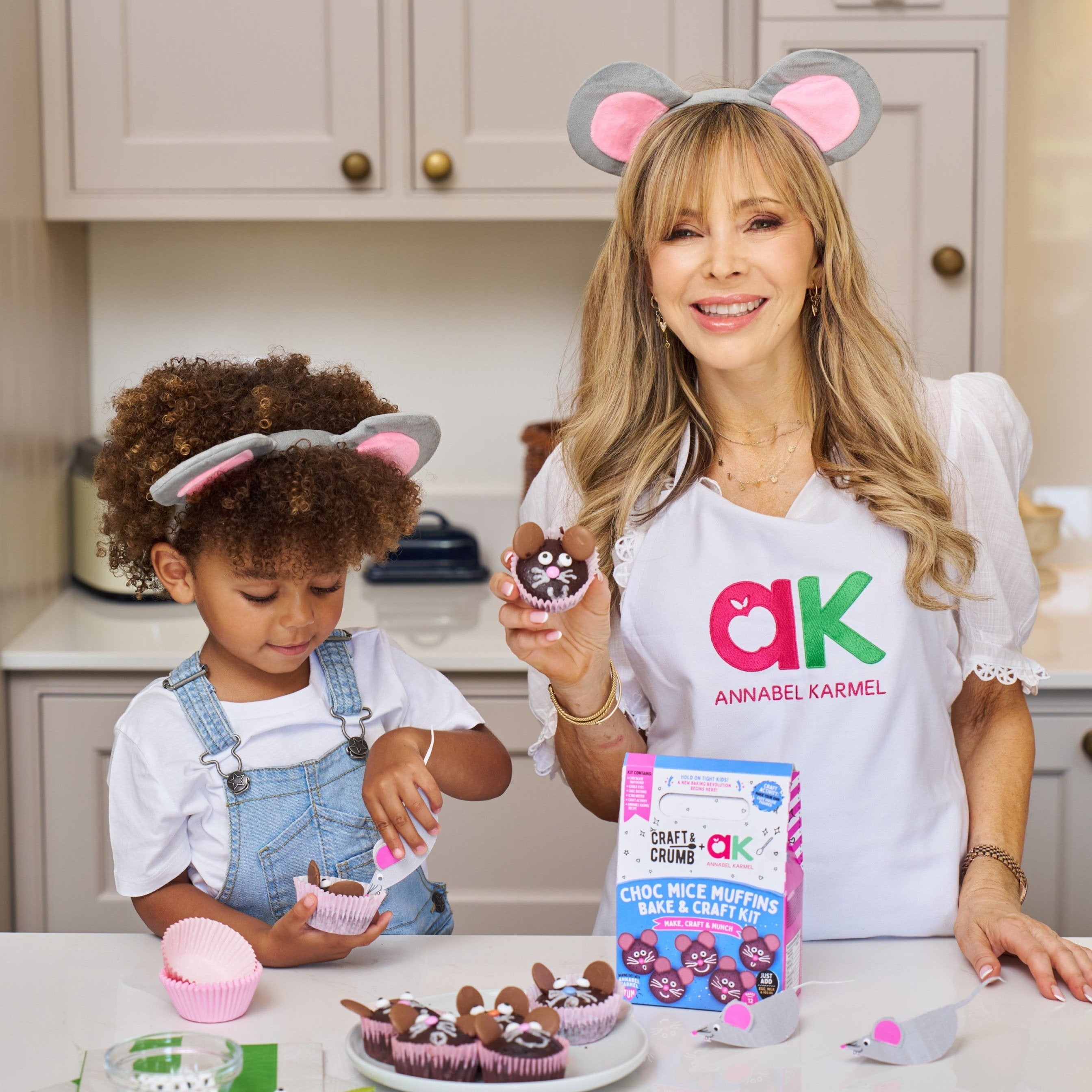 Woman and child wearing mouse ears baking chocolate mouse-themed muffins in a kitchen.