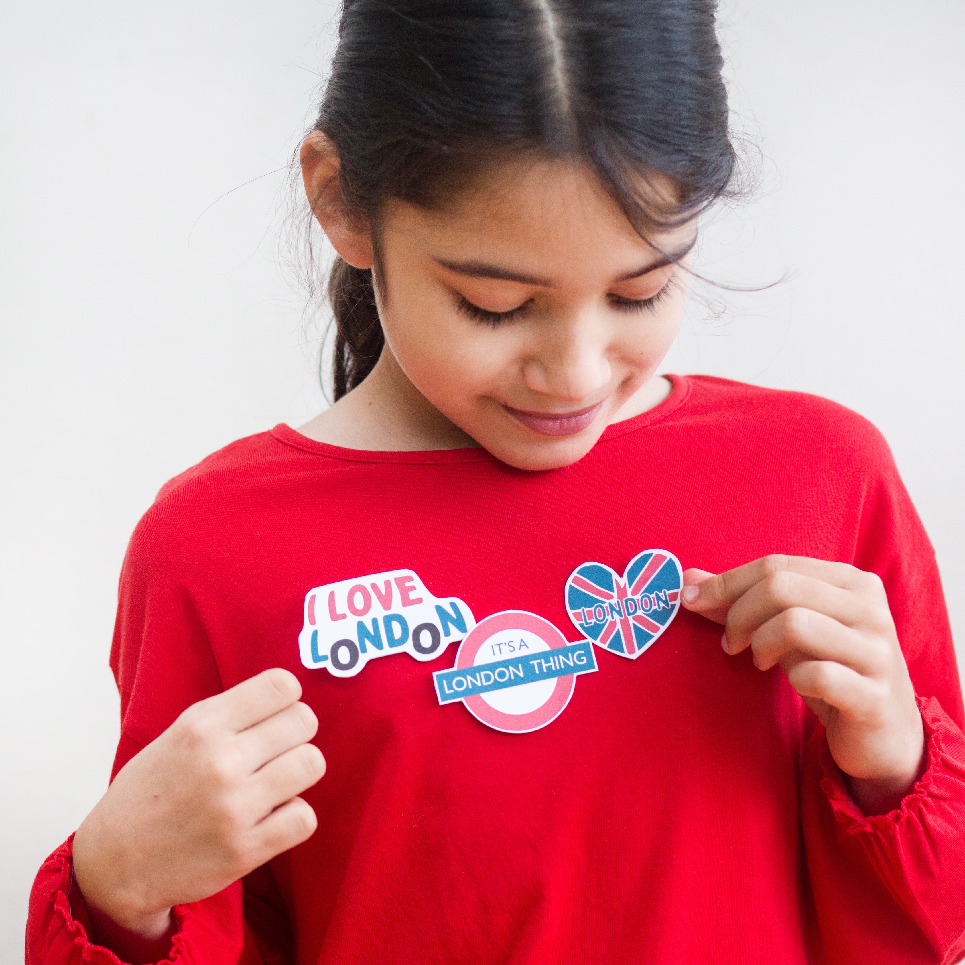 Girl in red shirt looking at London-themed stickers on her chest, including "I Love London" and a Union Jack heart.