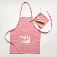Red and white striped apron with "Craft & Crumb" text, matching pouch, and wooden spoon on a light background.