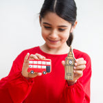 Girl in red holding cookies shaped like a red bus and Big Ben, smiling against a plain background.