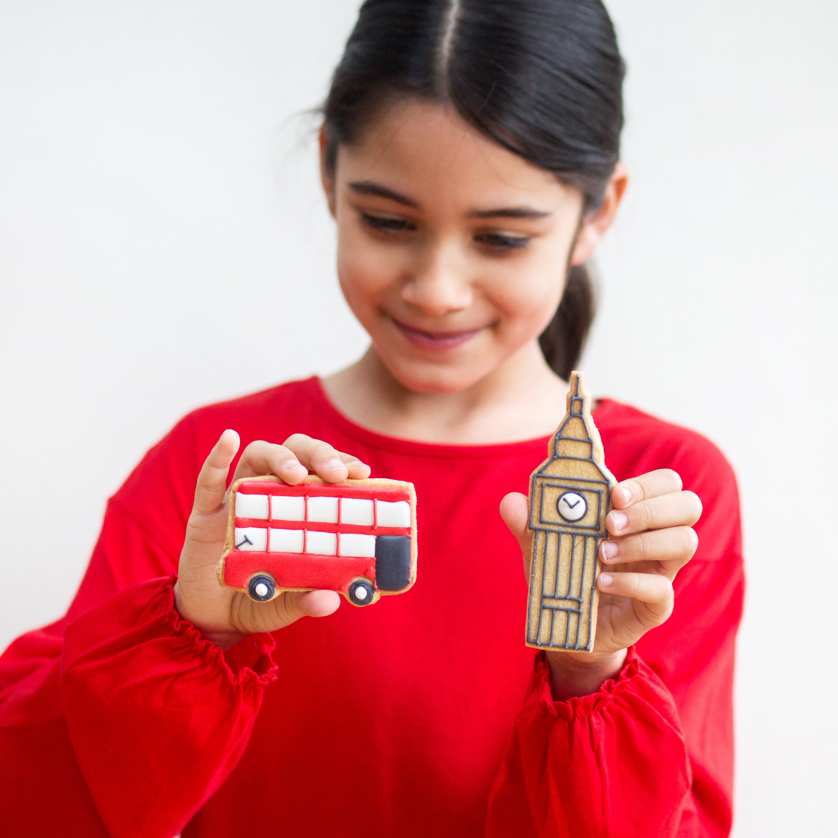 Girl in red holding cookies shaped like a red bus and Big Ben, smiling against a plain background.