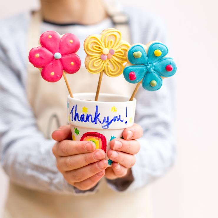 Child holding a decorated cup with colorful flower-shaped cookies on sticks, featuring a "Thank you!" message, from Craft & Crumb's baking kits for kids.