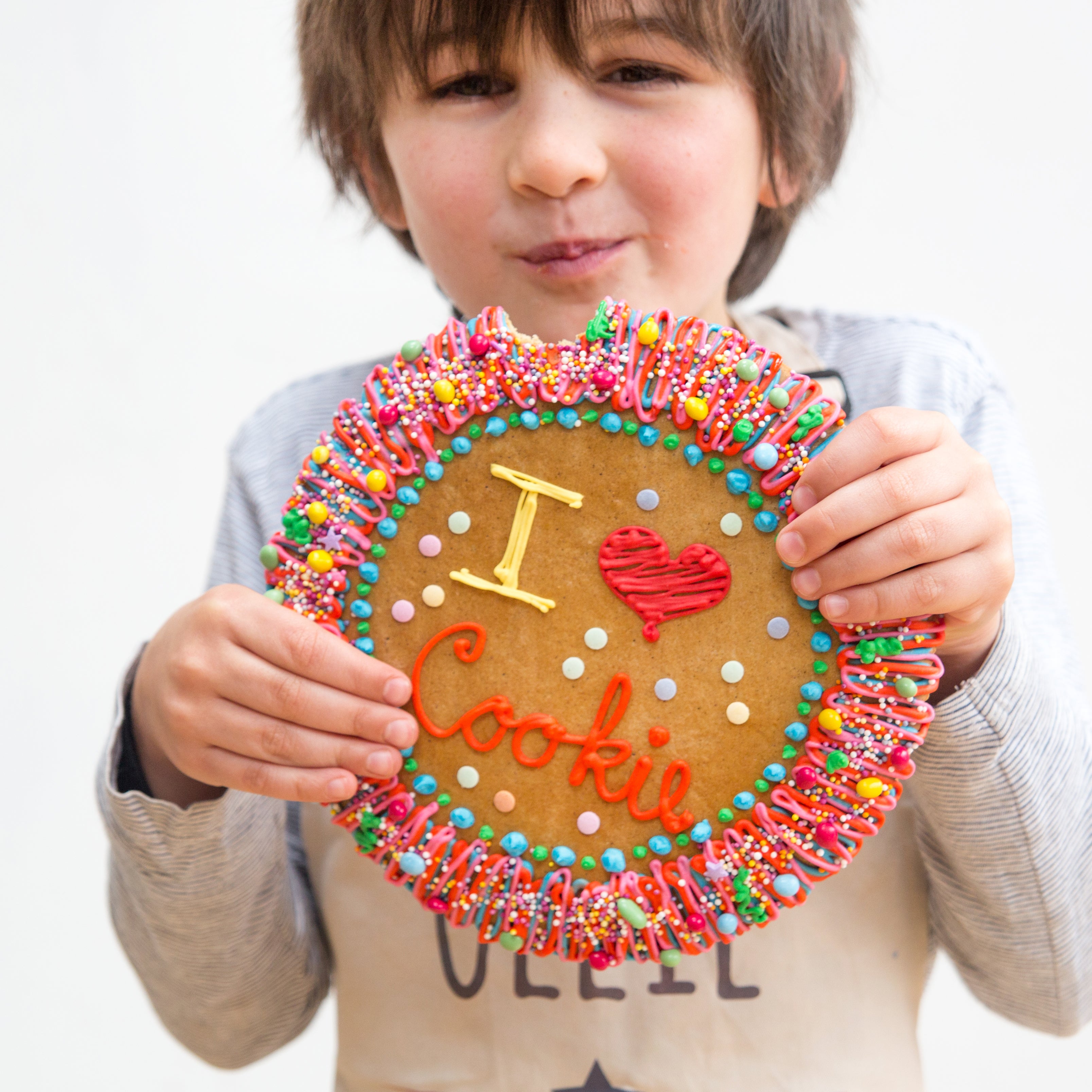 Child holding a large decorated cookie with colorful icing and sprinkles, featuring the text "I ♥ Cookie," from Craft & Crumb's baking kits for kids in the UK.