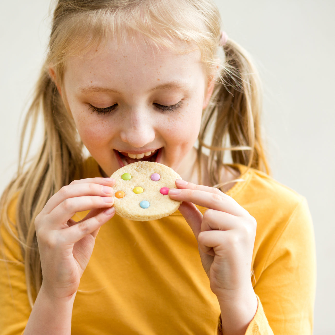Girl in yellow shirt eating a cookie with colorful candy toppings, smiling and looking down at the cookie.