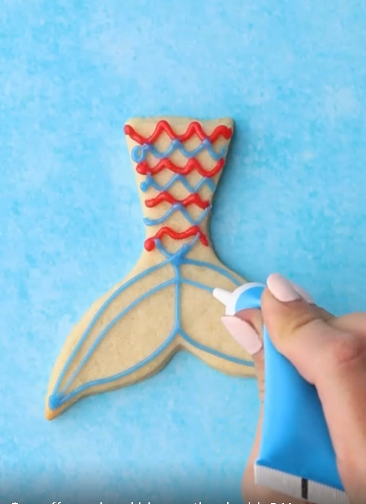 Hand decorating a mermaid tail-shaped cookie with blue and red icing on a blue background.