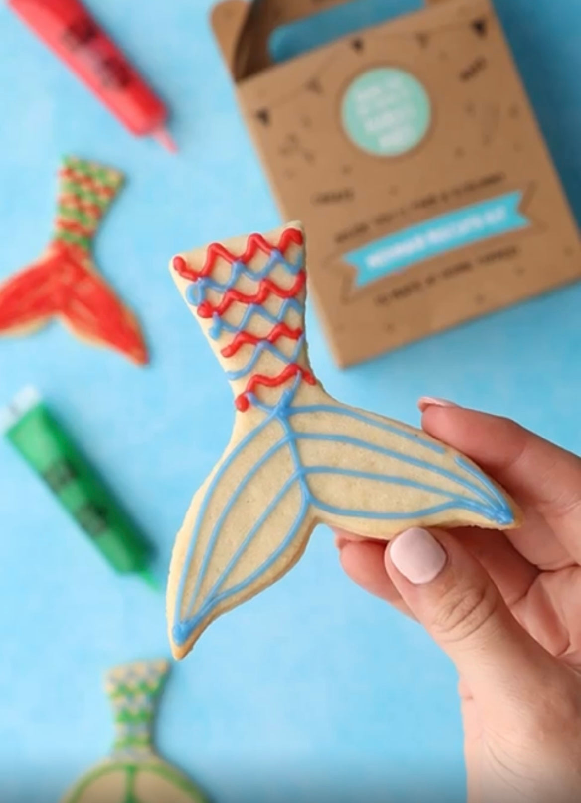 Hand holding a decorated mermaid tail cookie with icing tubes and a cookie kit box in the background.
