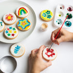 Person decorating cookies with colorful designs and paintbrush, surrounded by painted cookies and paint palette.