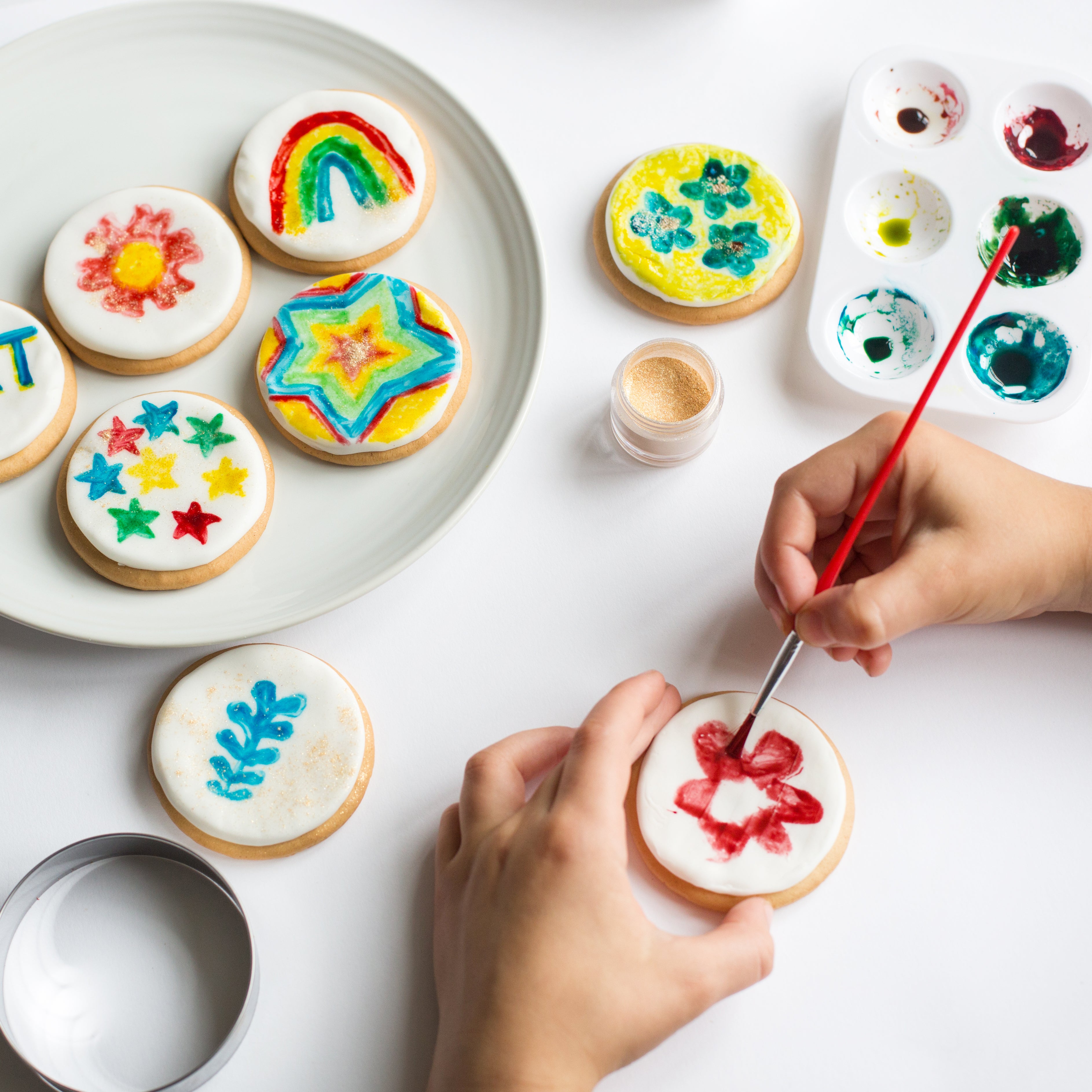 Person decorating cookies with colorful designs and paintbrush, surrounded by painted cookies and paint palette.