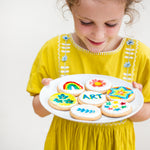 Child in yellow dress holding a plate of decorated cookies with colorful designs and the word "ART."