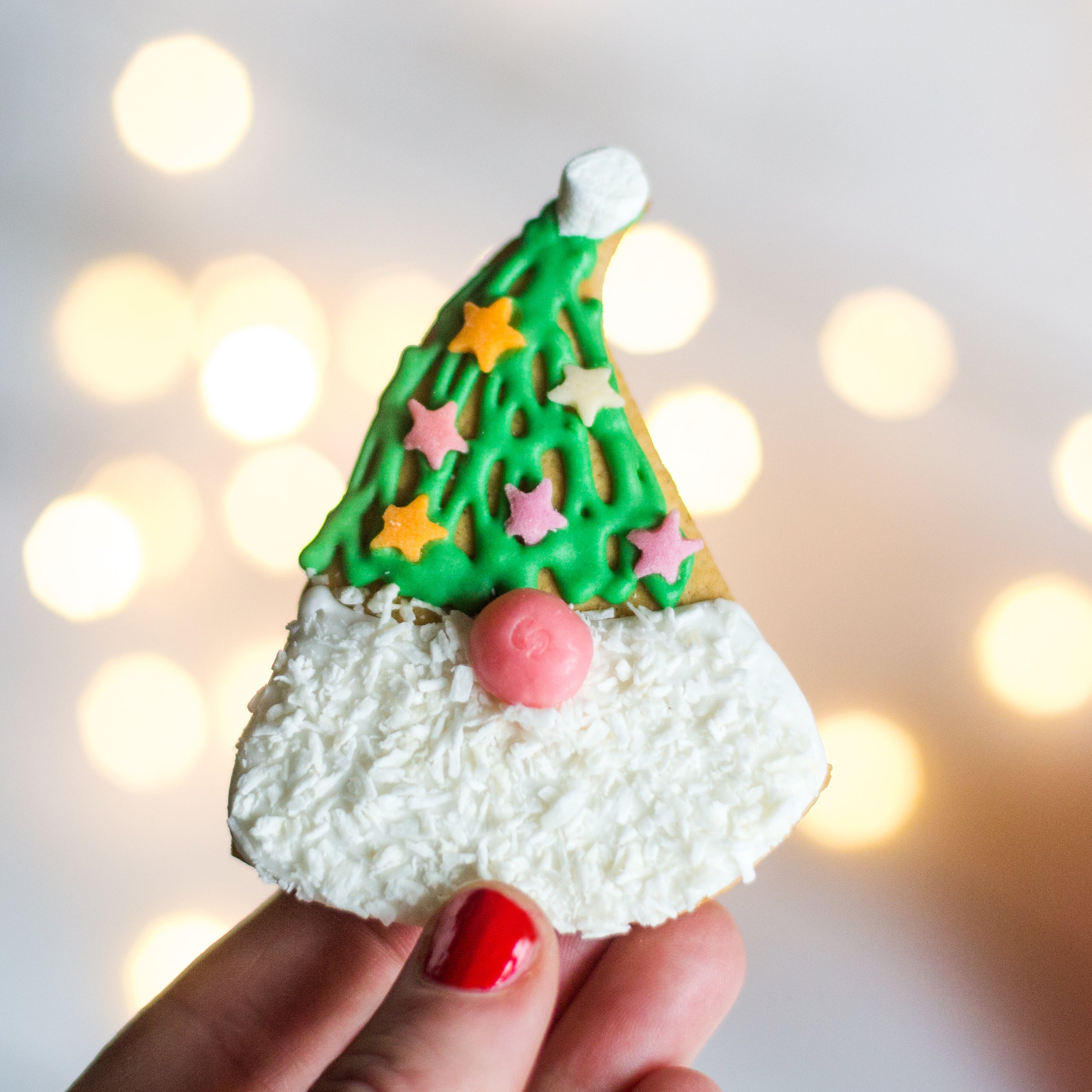 Hand holding a gnome-shaped cookie with green hat, star sprinkles, and white icing beard, against blurred lights background.