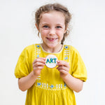 Girl in a yellow dress holding a cookie with "ART" written on it, smiling against a plain background.