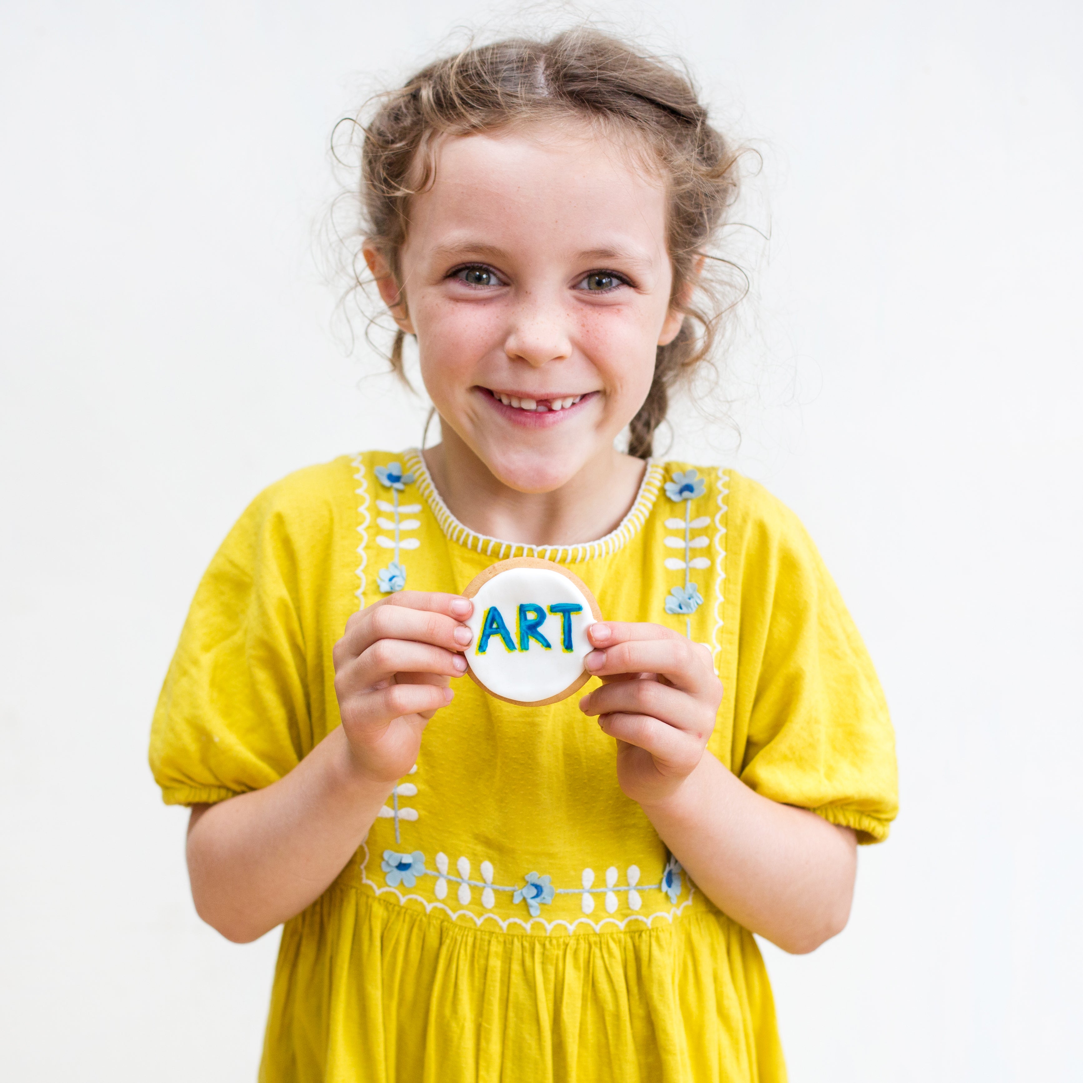 Girl in a yellow dress holding a cookie with "ART" written on it, smiling against a plain background.