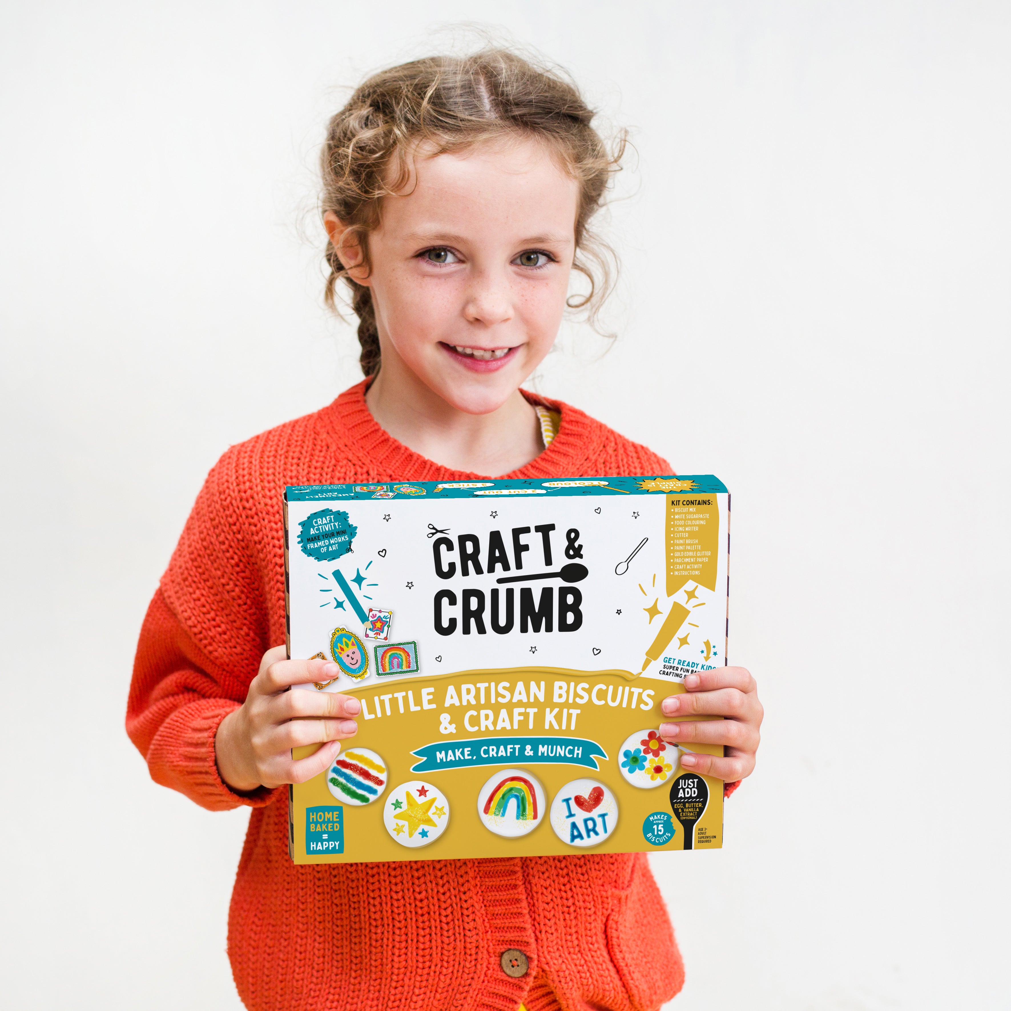 Child holding a "Craft & Crumb" biscuit and craft kit, wearing an orange sweater, smiling at the camera.