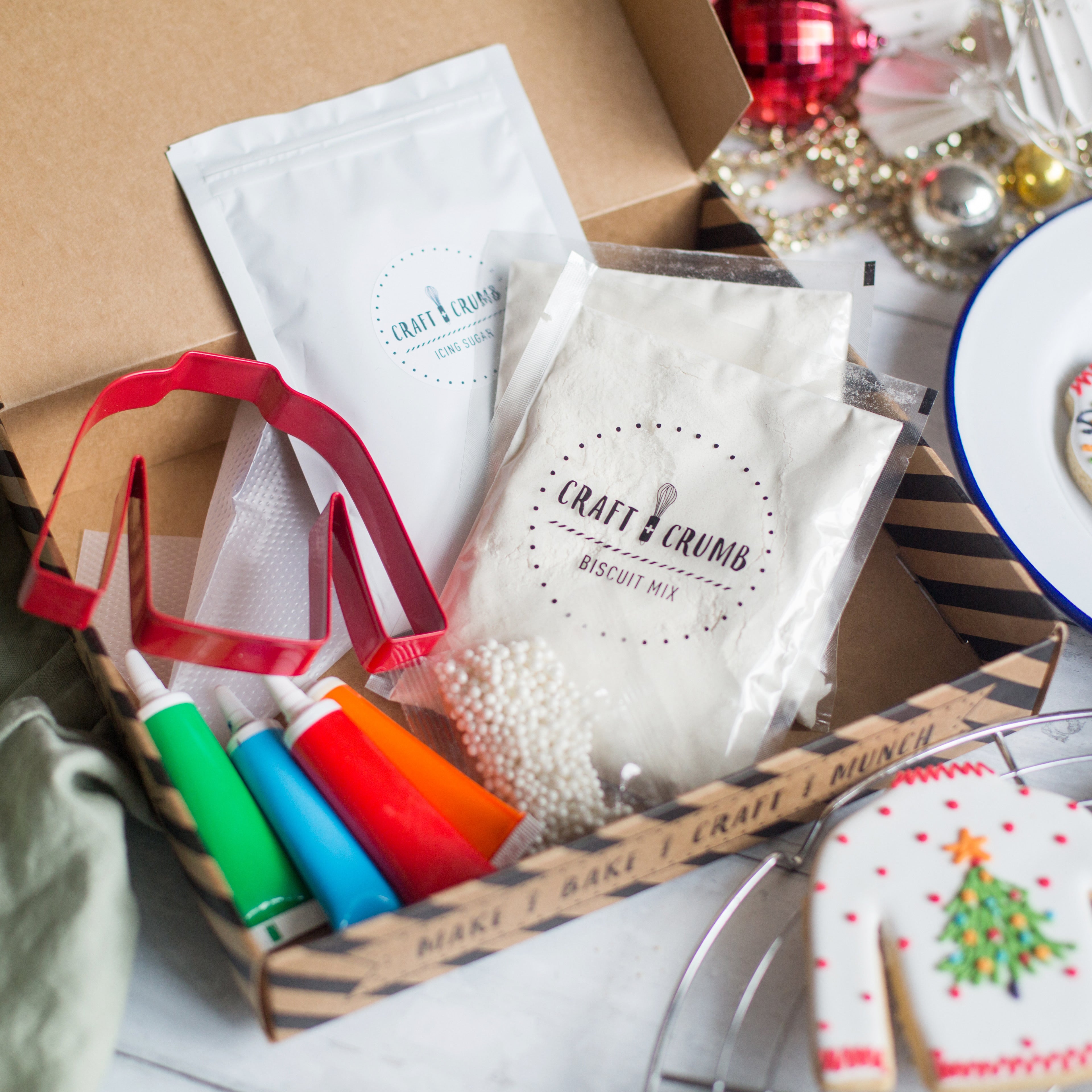 Baking kit from Craft & Crumb featuring biscuit mix, icing sugar, colorful icing tubes, a red cookie cutter, and decorative sprinkles, ideal for kids' baking activities.
