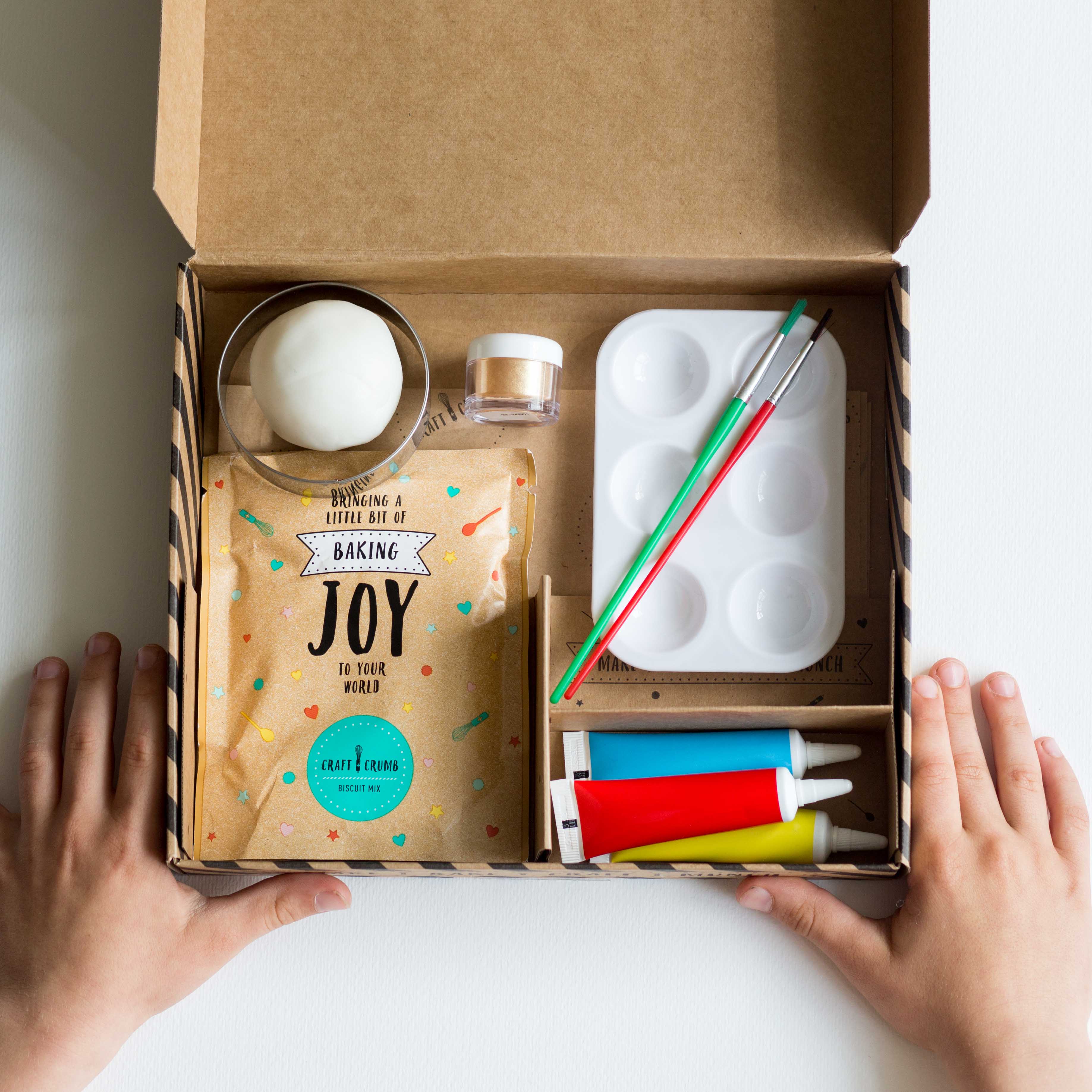 Baking kit with colorful icing tubes, brushes, palette, and dough in an open box, hands holding the sides.