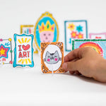 Hand holding a small, colorful drawing of a cat in a frame, surrounded by other mini art pieces on stands.
