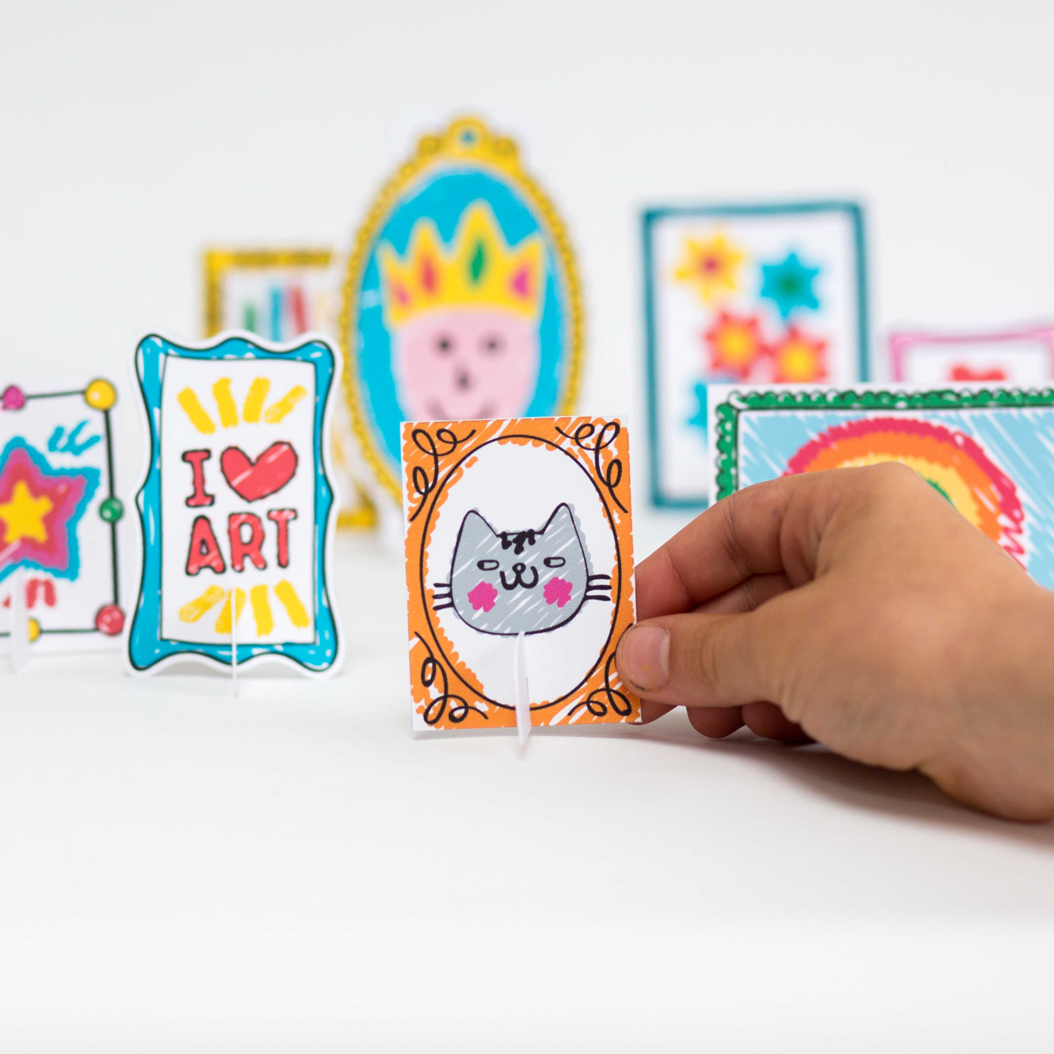 Hand holding a small, colorful drawing of a cat in a frame, surrounded by other mini art pieces on stands.