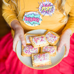 Child holding a plate of frosted pastries with colorful sprinkles, wearing a yellow shirt and pink skirt with comic-style stickers.