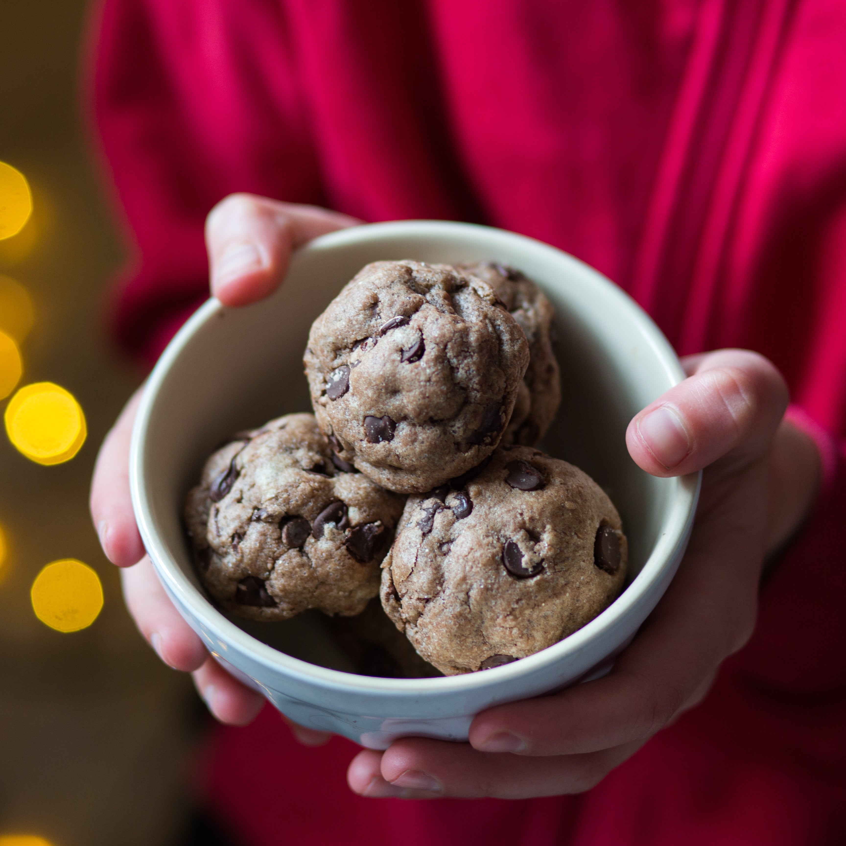 Hands holding a bowl of chocolate chip cookies, with a blurred background of warm yellow lights.