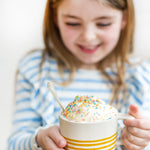 Child holding a mug with ice cream topped with colorful sprinkles, wearing a striped shirt, from Craft & Crumb's baking kits for kids.