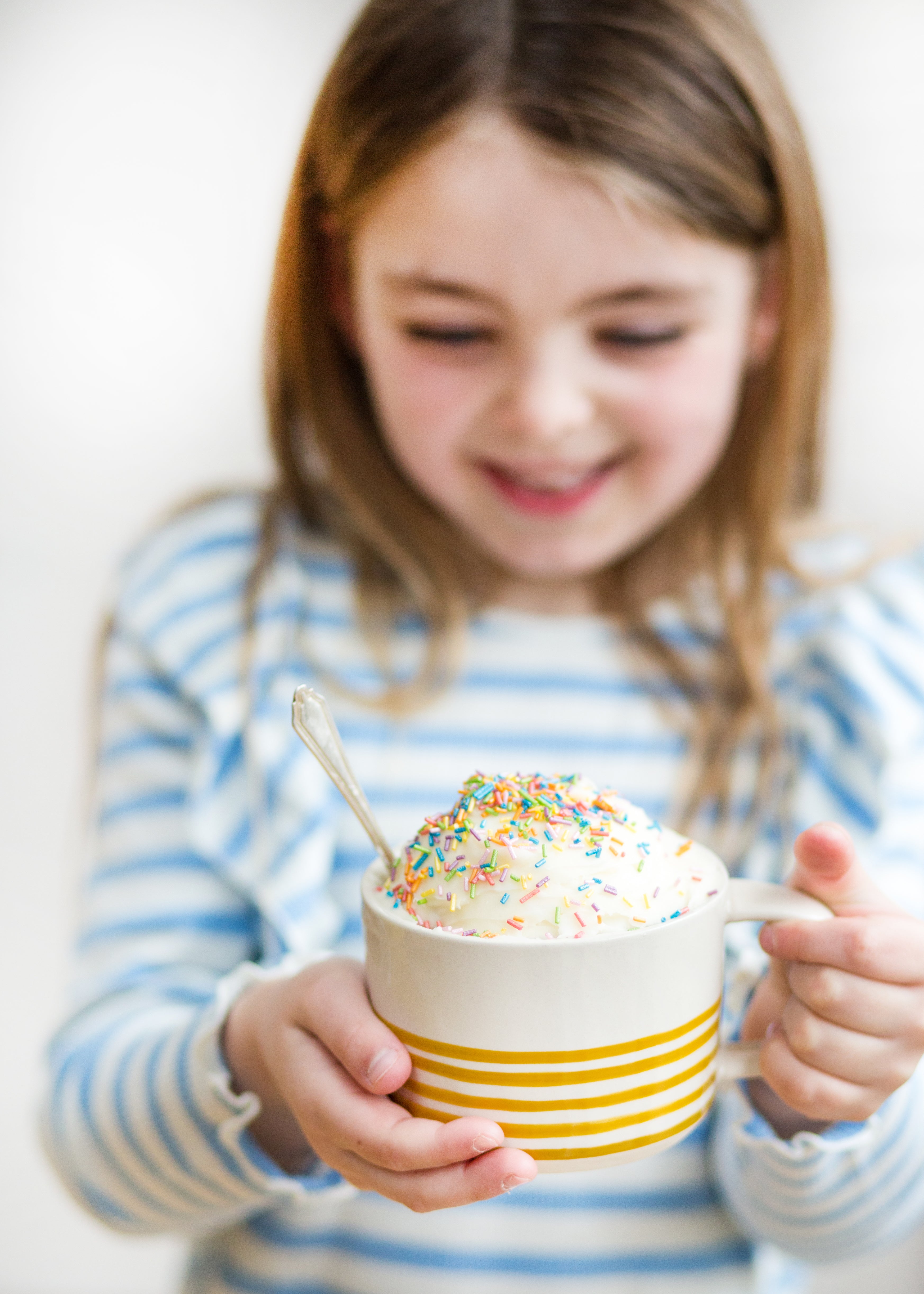 Child holding a mug with ice cream topped with colorful sprinkles, wearing a striped shirt, from Craft & Crumb's baking kits for kids.