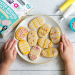 Child decorating Easter egg-shaped cookies with icing and sprinkles on a plate.