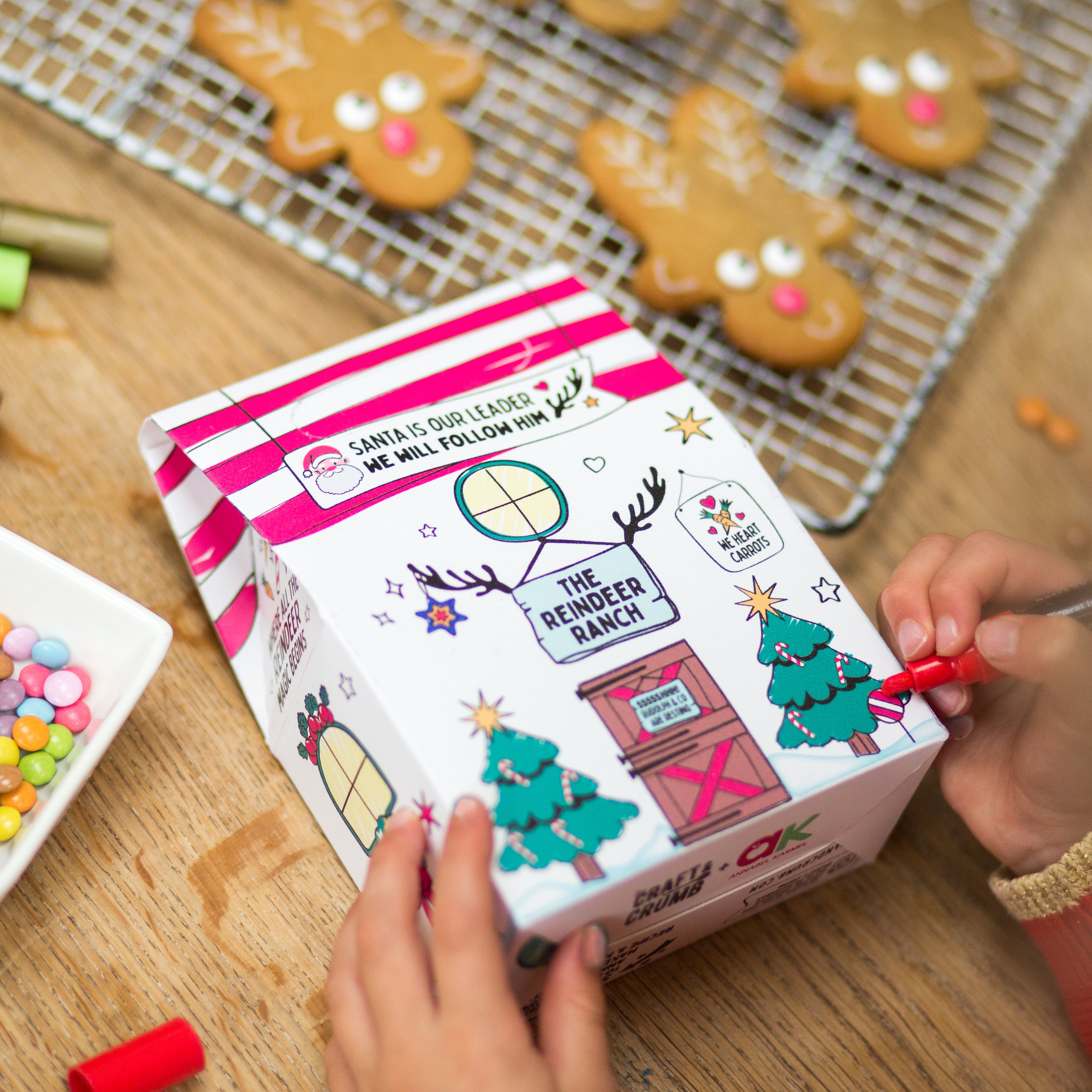 Child decorating a festive box with reindeer cookies and colorful candies on a wooden table.