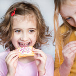 Child with curly hair eating a sprinkled pastry, wearing a pink shirt, next to another child in a yellow shirt.