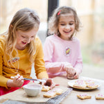 Two young girls decorating cookies with icing and sprinkles at a table, smiling and enjoying the activity.