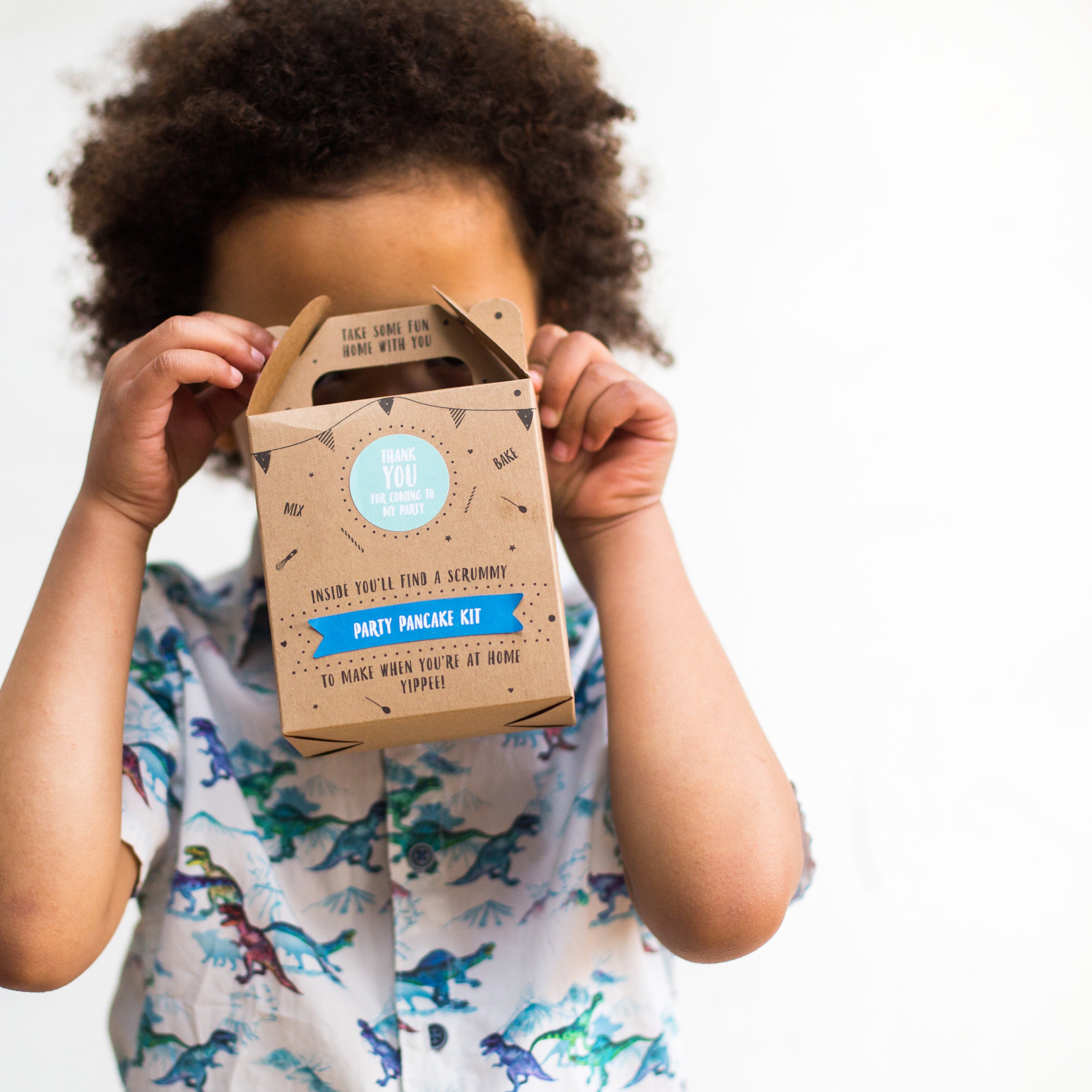 Child holding a party pancake kit box, wearing a dinosaur shirt, against a white background.