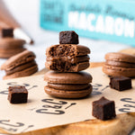 Chocolate macarons stacked with brownie pieces on parchment paper, with a macaron box in the background.