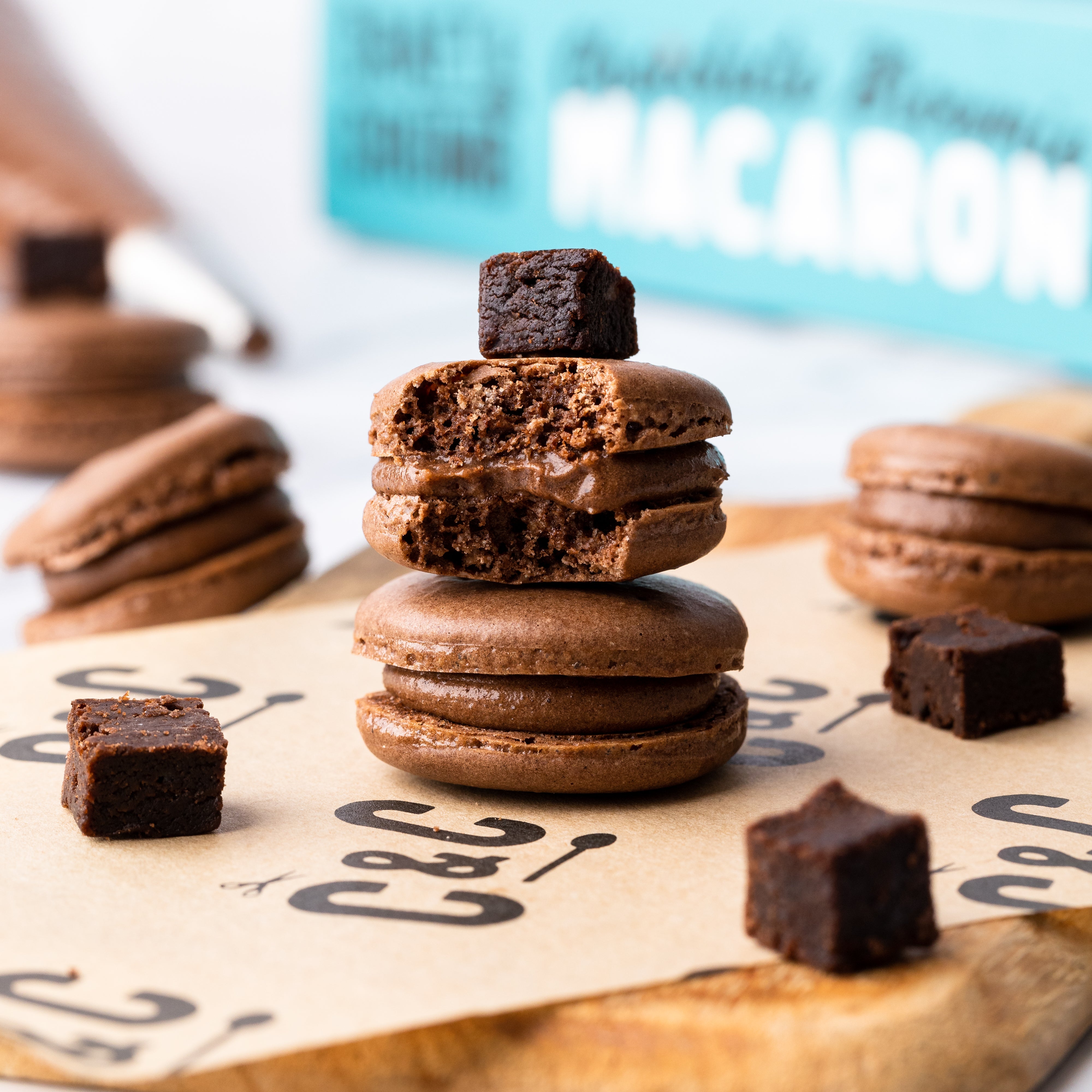 Chocolate macarons stacked with brownie pieces on parchment paper, with a macaron box in the background.