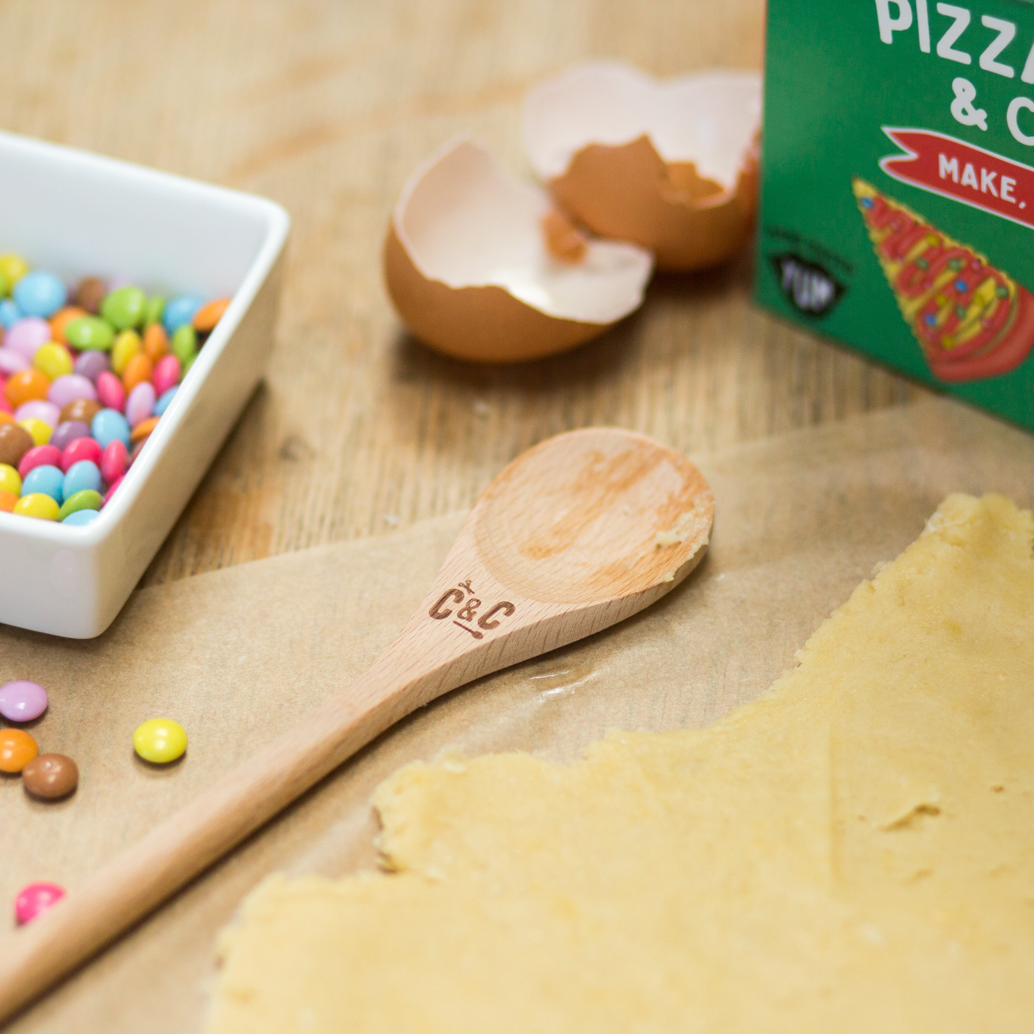 Wooden spoon, cookie dough, colorful candies, eggshells, and pizza box on a wooden surface.