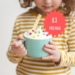 Child holding a turquoise mug with whipped cream and sprinkles, wearing a striped shirt. Price tag reads "£3 per bag."