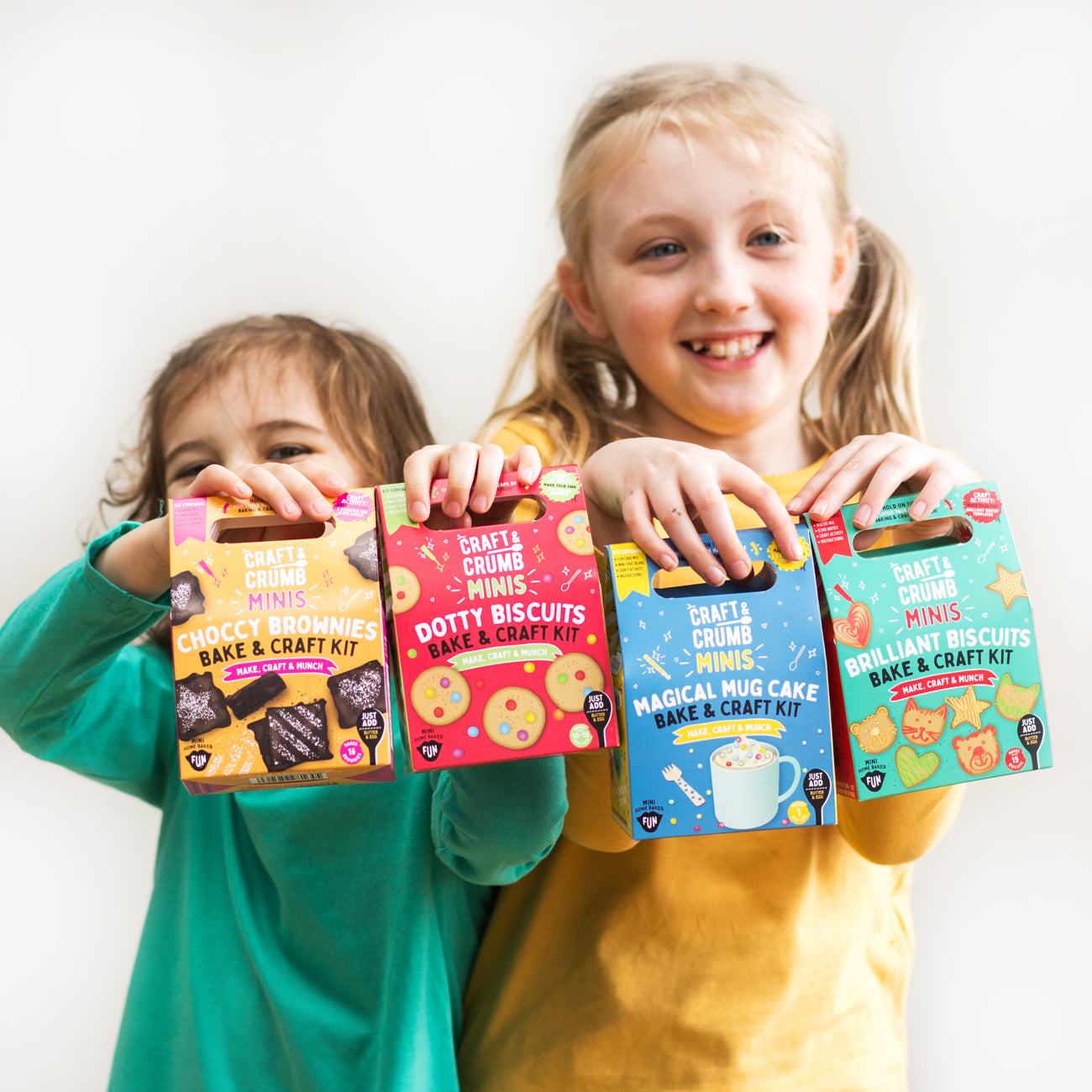 Two children smiling and holding colorful Craft & Crumb baking kits, including brownies, biscuits, and mug cake.