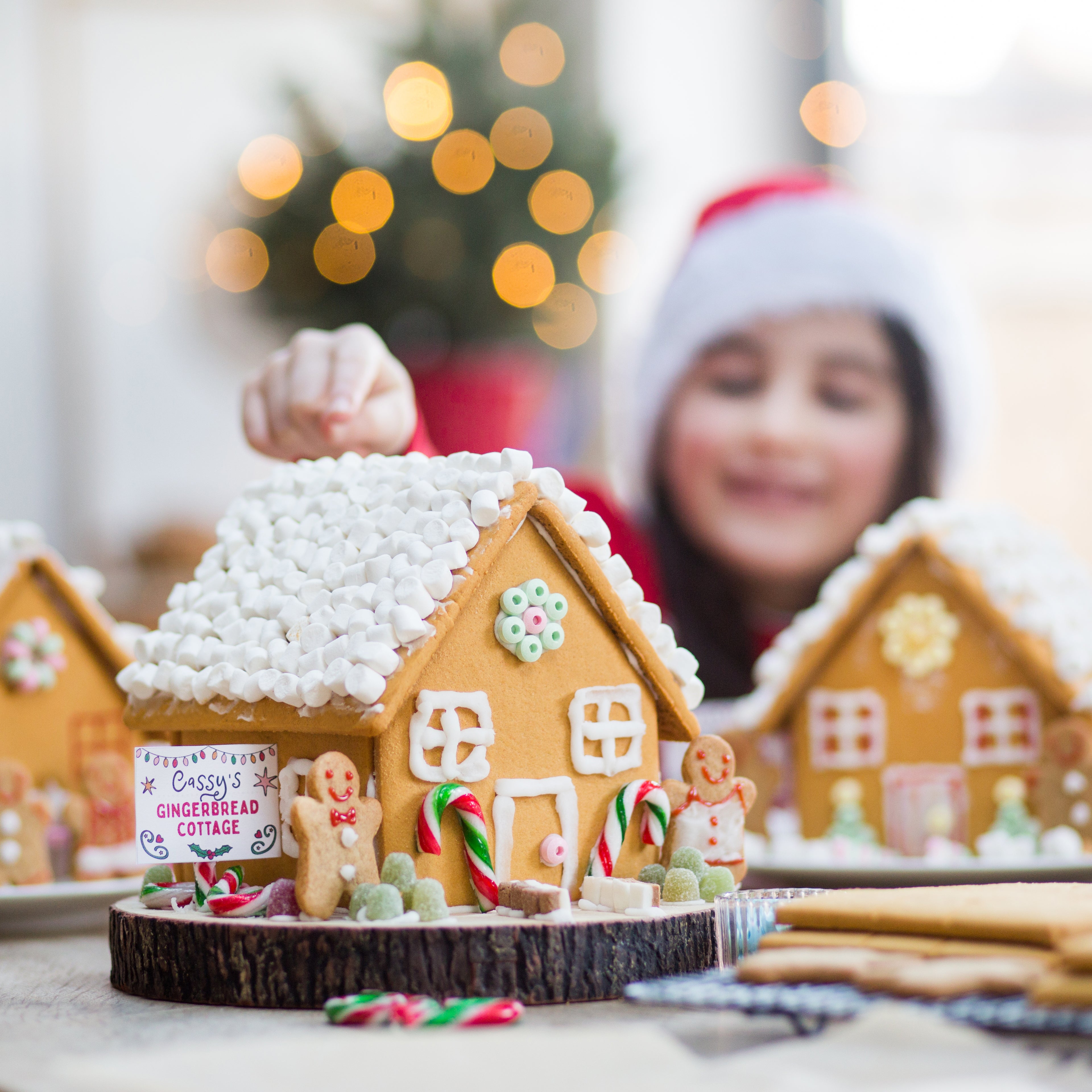 Child decorating gingerbread house with candy, wearing Santa hat, blurred festive lights in background.