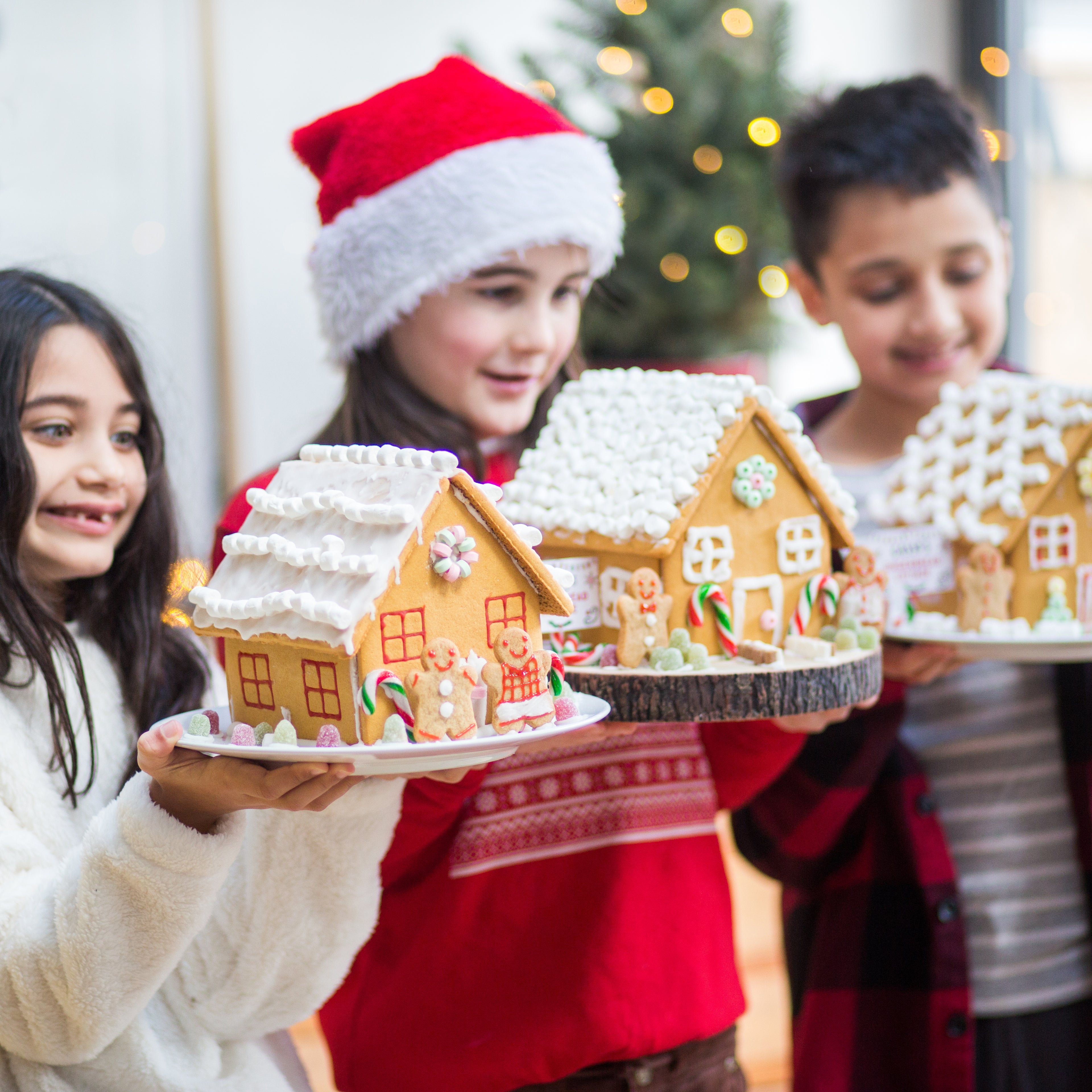 Three children holding decorated gingerbread houses, with one wearing a Santa hat, in a festive setting.