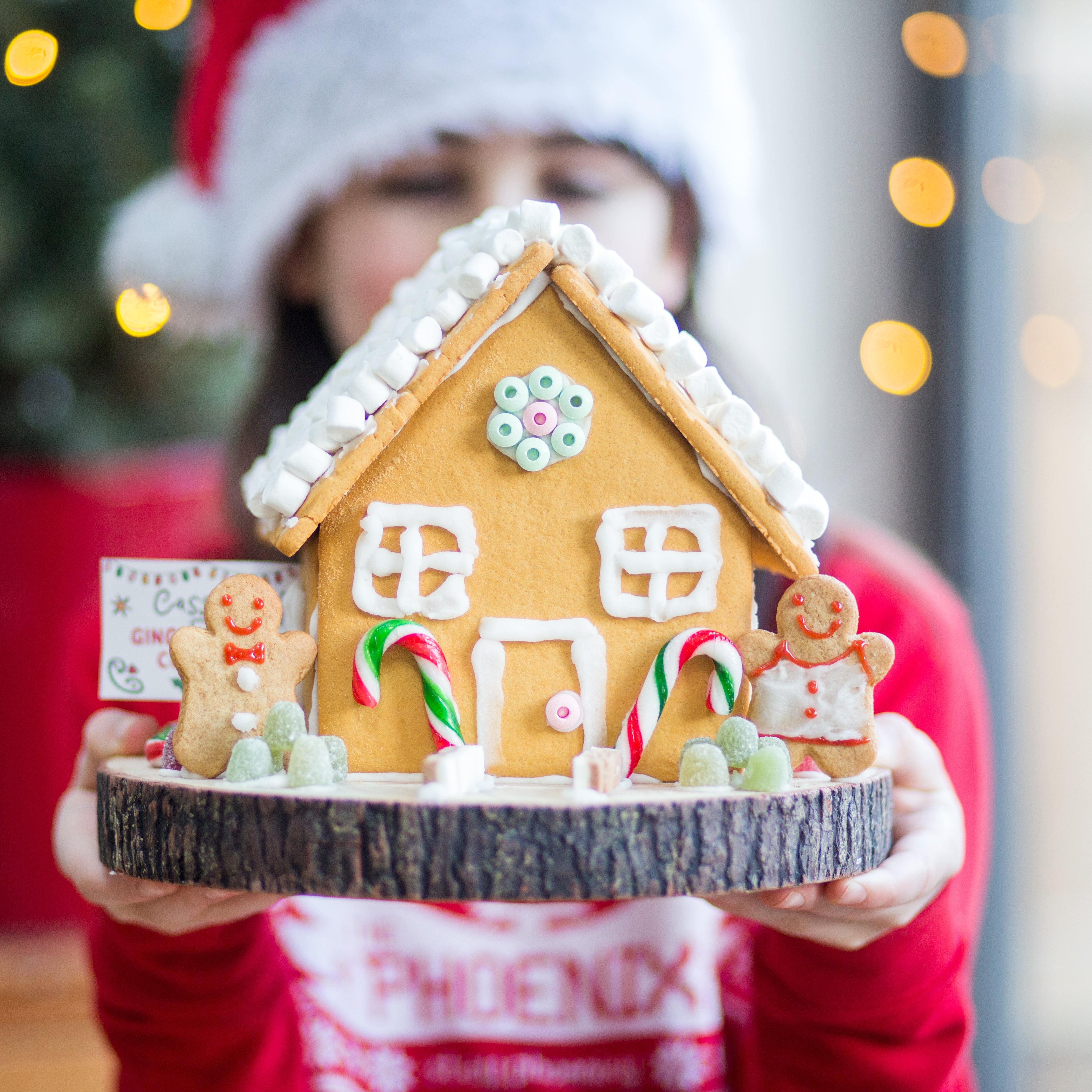 Person holding a decorated gingerbread house with candy canes and gingerbread figures, wearing a Santa hat.