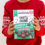 Child holding a "Reindeer Poops Cookies & Craft Kit" box, wearing a red sweater with festive patterns.