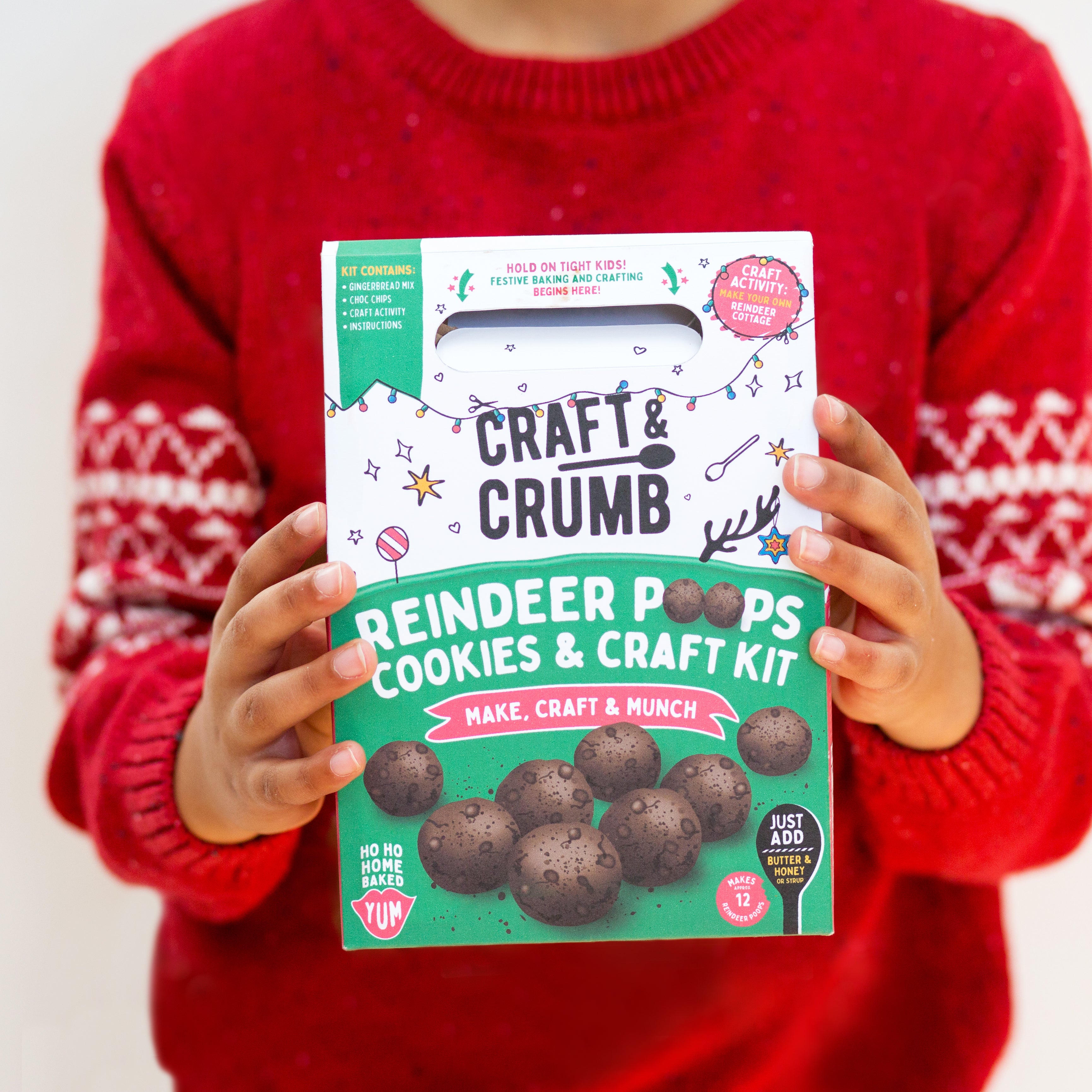 Child holding a "Reindeer Poops Cookies & Craft Kit" box, wearing a red sweater with festive patterns.