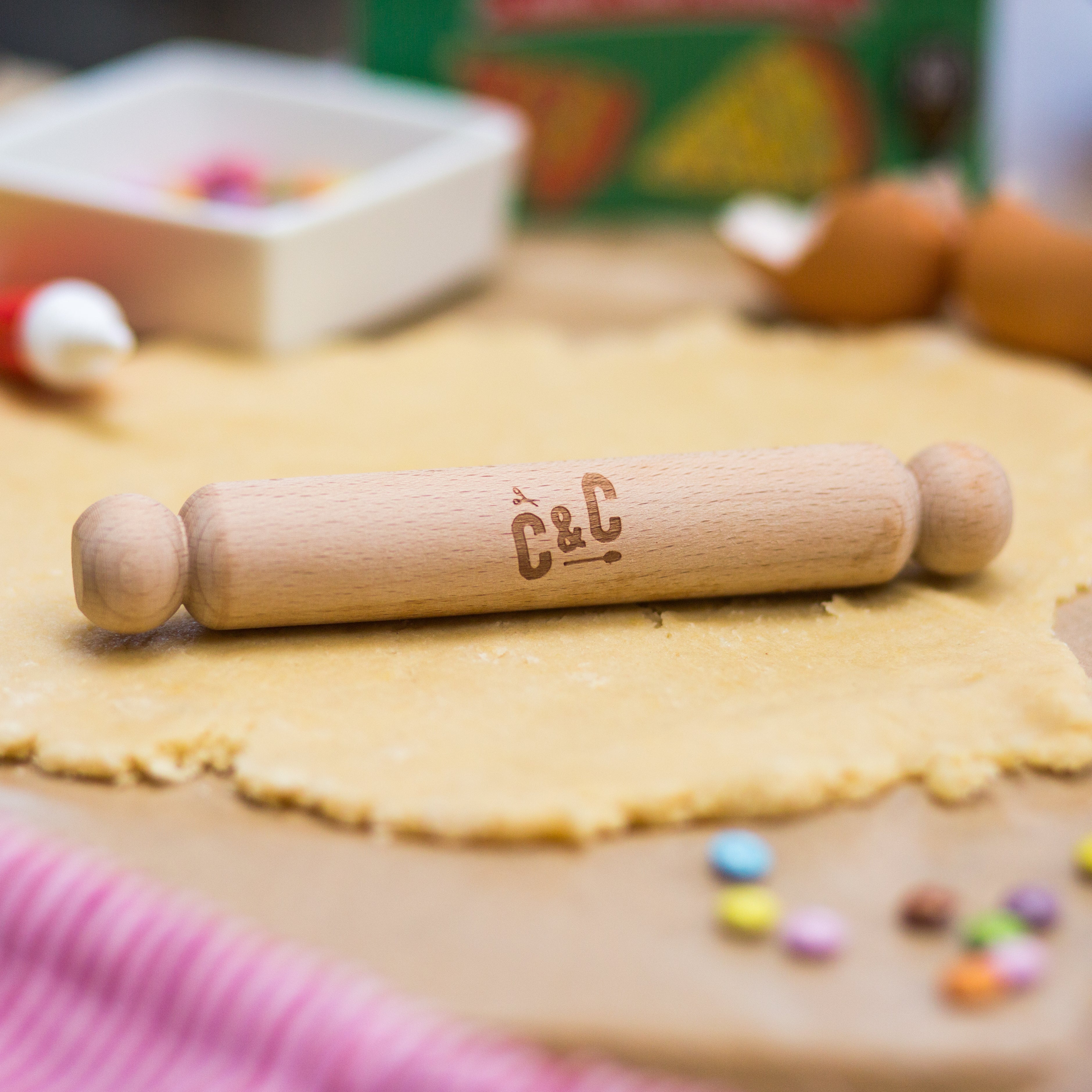 Wooden rolling pin on dough with colorful candies and baking items in the background.