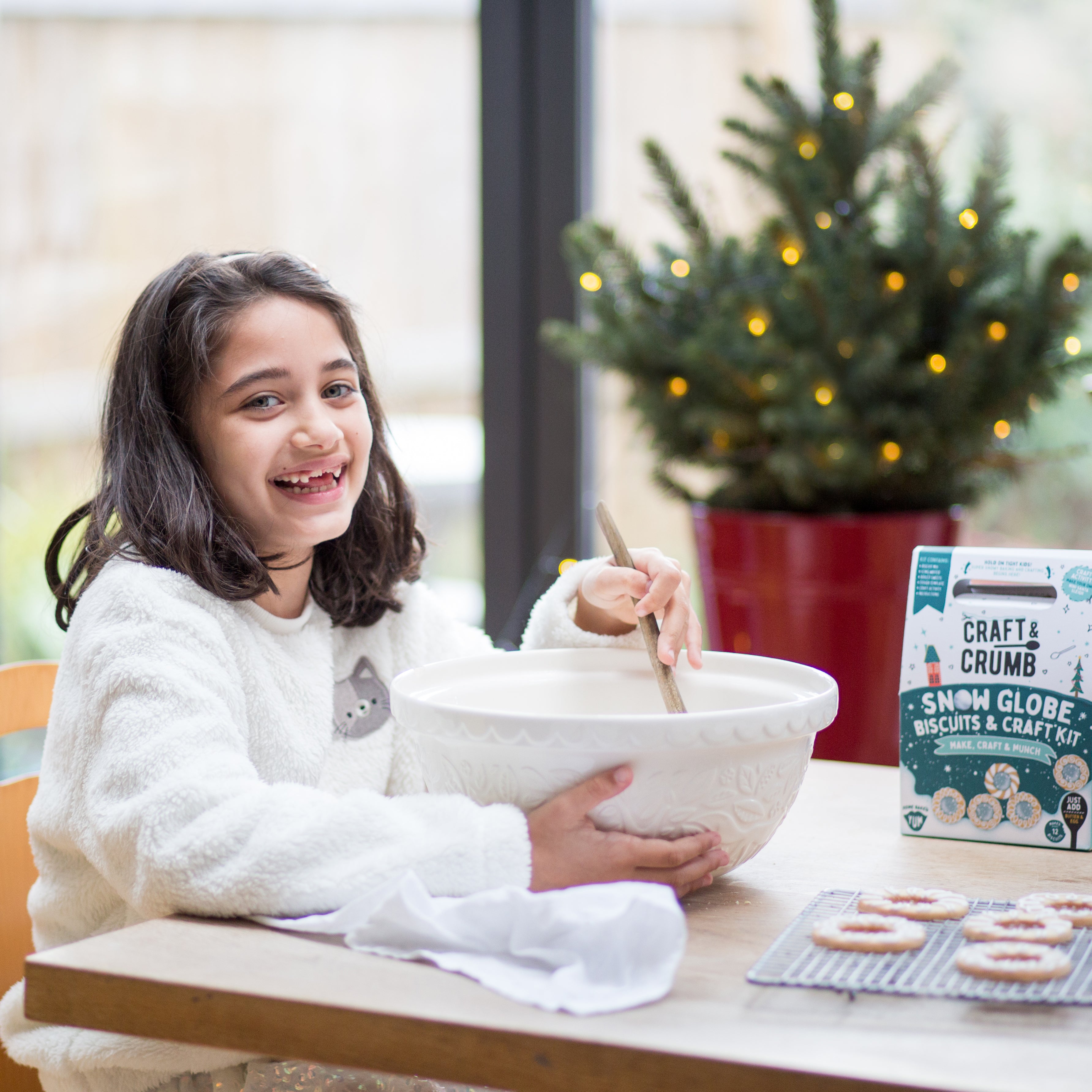 Girl smiling while mixing dough in a bowl, with a small decorated Christmas tree and biscuit kit on the table.