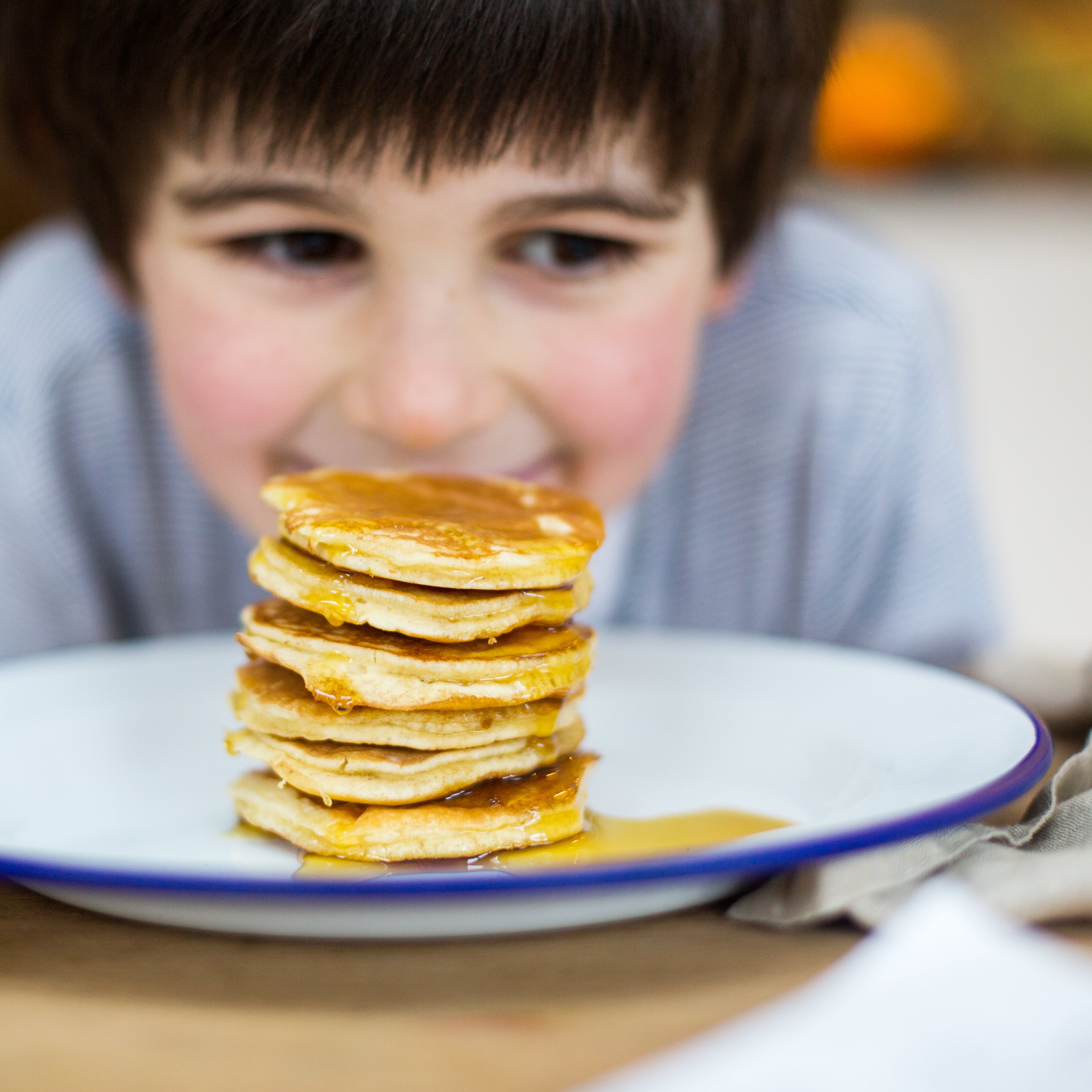 Child smiling at a stack of pancakes with syrup on a white plate.