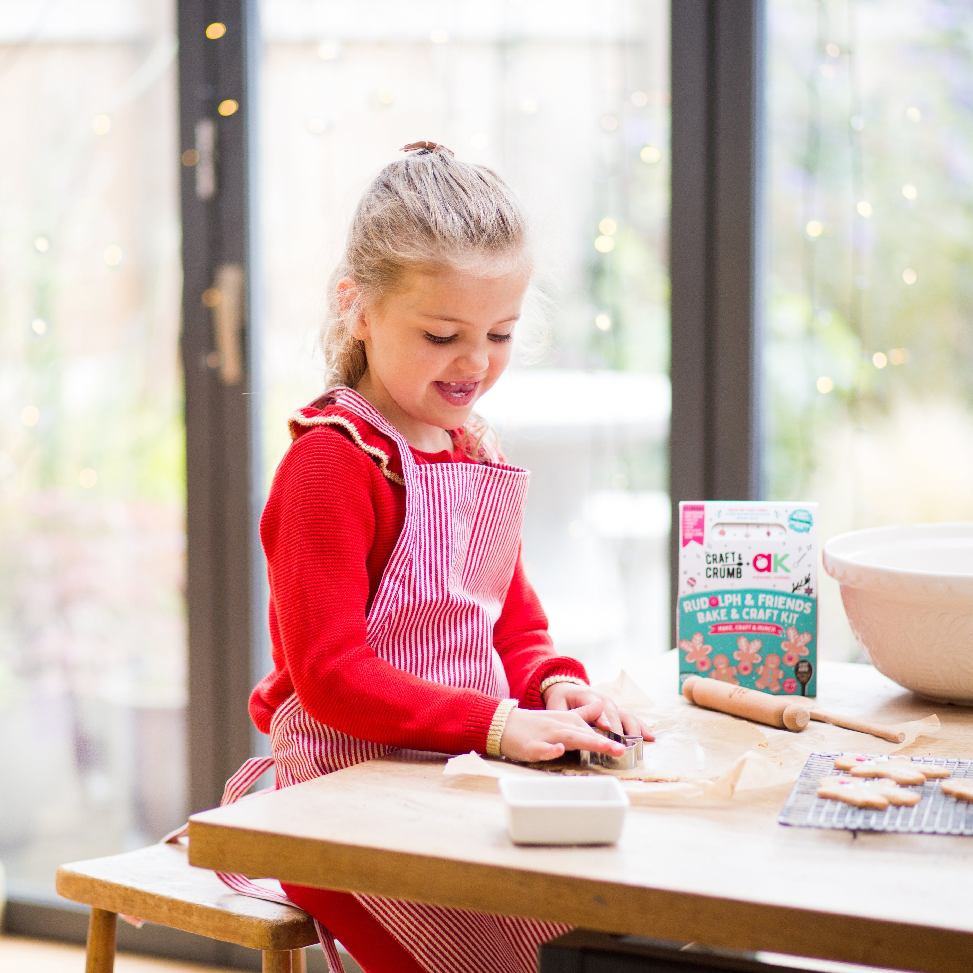 Young girl in red apron making cookies at a kitchen table, with a craft kit and bowl nearby.