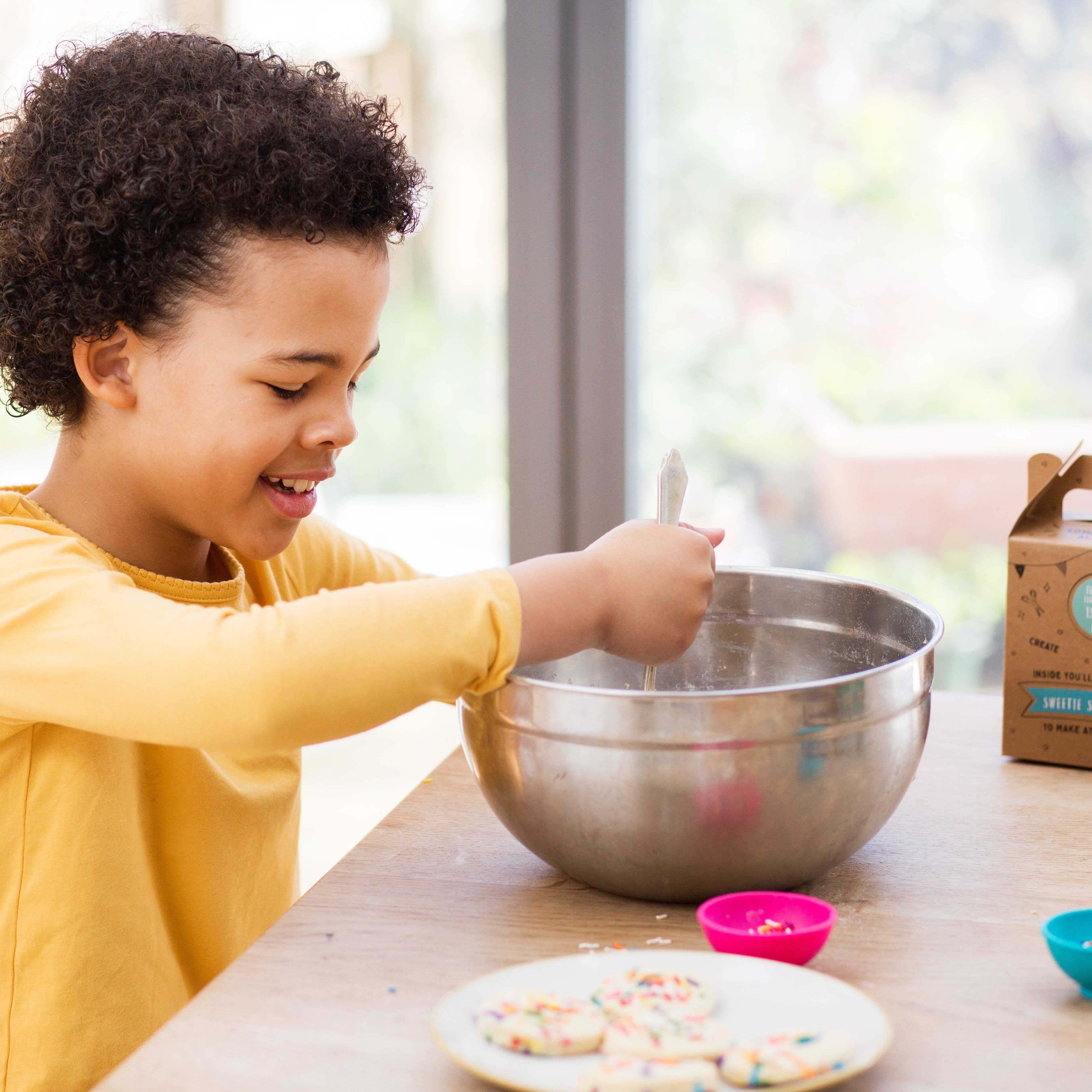 Child mixing batter in a metal bowl at a table with decorated cookies and baking supplies.