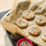 Cookies with colorful sprinkles on parchment paper, next to a pink bowl of extra sprinkles.