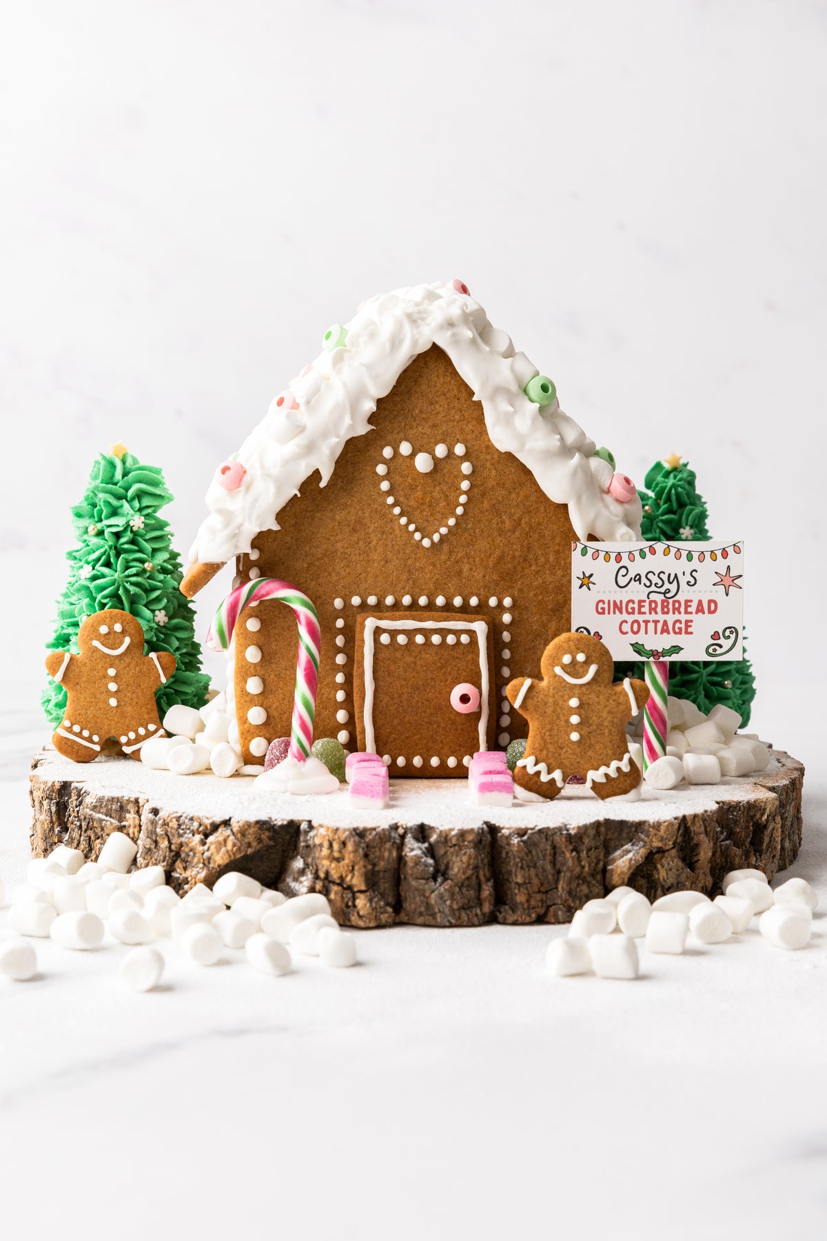 Gingerbread house with icing, candy decorations, and gingerbread figures on a wooden base, surrounded by mini marshmallows, from Craft & Crumb's baking kits for kids.